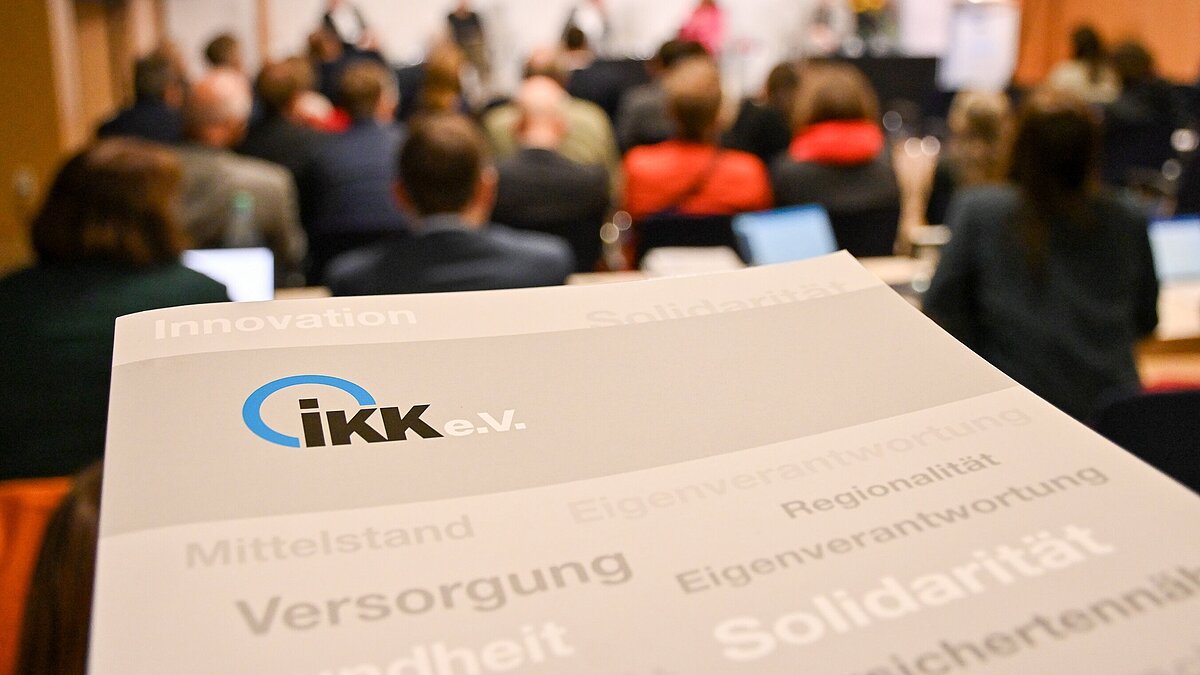 Pressemappe des IKK e.V. im Vordergrund, im Hintergrund Menschen auf einer Tagung